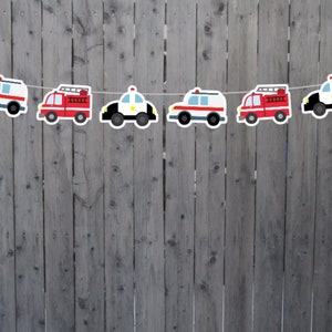 May include: A string of six paper cutouts of vehicles, alternating between red fire trucks and white police cars. The vehicles are strung together with white string and are hanging against a gray wooden fence.