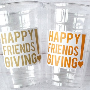 May include: Two clear plastic cups with the text "HAPPY FRIENDS GIVING" in gold and orange, with a heart symbol. The cups are empty and ready for beverages.