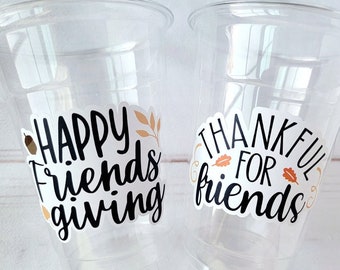 May include: Two clear plastic disposable cups featuring adhesive decals. The left cup displays the black text "HAPPY Friends giving" with a small brown acorn and orange leaf. The right cup shows the black text "THANKFUL FOR friends" with orange leaves.