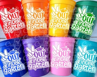 Puede incluir: Conjunto de diez vasos de plástico de colores con una calcomanía de vinilo blanco que dice "OUT kinder garten" con un signo de paz y un gorro de graduación.