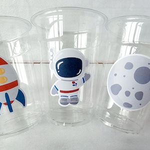 Op de afbeelding: Drie doorzichtige plastic bekers met ruimtethema stickers. E&eacute;n beker heeft een raket, &eacute;&eacute;n heeft een astronaut en &eacute;&eacute;n heeft een maan.