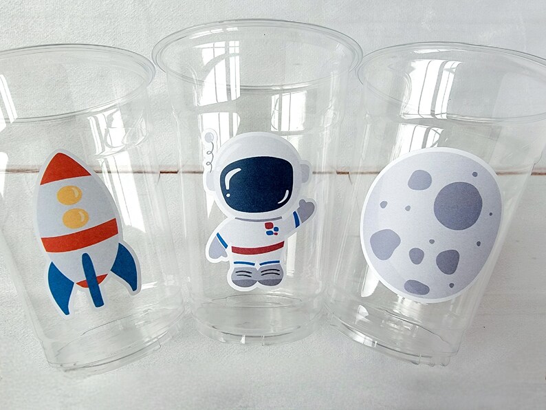 Op de afbeelding: Drie doorzichtige plastic bekers met ruimtethema stickers. E&eacute;n beker heeft een raket, &eacute;&eacute;n heeft een astronaut en &eacute;&eacute;n heeft een maan.