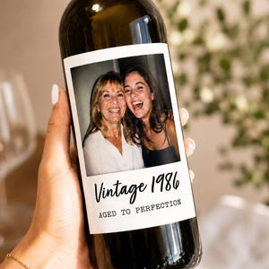 May include: A dark glass wine bottle featuring a custom rectangular label with a photograph of two smiling individuals. The label includes the black script text Vintage 1986 and the block text AGED TO PERFECTION printed below the photo.