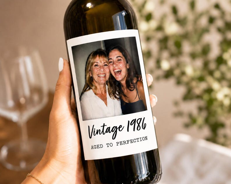 May include: A dark glass wine bottle featuring a custom rectangular label with a photograph of two smiling individuals. The label includes the black script text Vintage 1986 and the block text AGED TO PERFECTION printed below the photo.