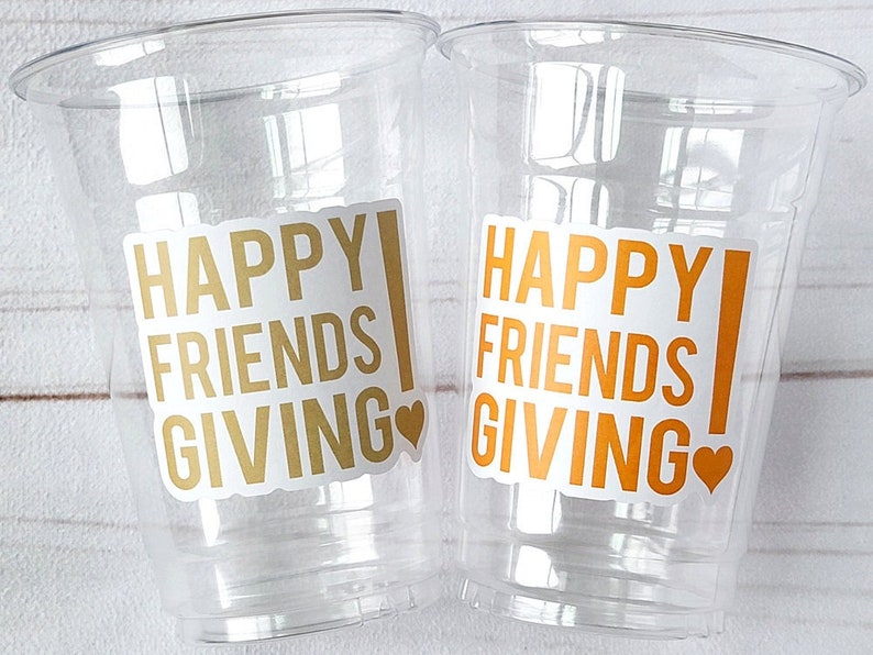 May include: Two clear plastic cups with the text "HAPPY FRIENDS GIVING" in gold and orange, with a heart symbol. The cups are empty and ready for beverages.