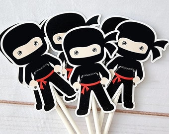 May include: Five black ninja cupcake toppers with red sashes and white eyes. Each topper has a black headband and is holding a black weapon.