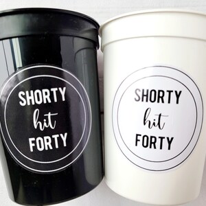 May include: Two plastic cups, one black and one white, with the text "SHORTY hit FORTY" on a circular sticker. The cups are likely for a party or event, with a celebratory theme.