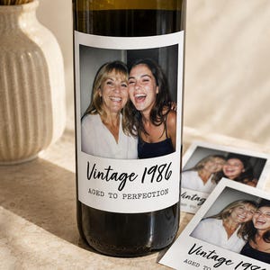May include: A dark glass wine bottle with a custom rectangular label featuring a photograph of two smiling individuals. The label includes the black printed text Vintage 1986 and AGED TO PERFECTION. Two matching printed cards are placed beside the bottle.