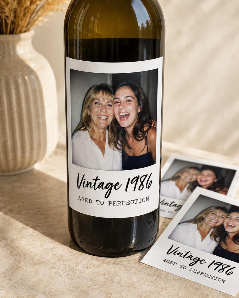 May include: A dark glass wine bottle with a custom rectangular label featuring a photograph of two smiling individuals. The label includes the black printed text Vintage 1986 and AGED TO PERFECTION. Two matching printed cards are placed beside the bottle.