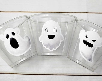 May include: Three clear plastic cups decorated with white ghost stickers. Each ghost has a different expression: one is smiling, one is screaming, and one is angry. The cups are arranged in a semi-circle.