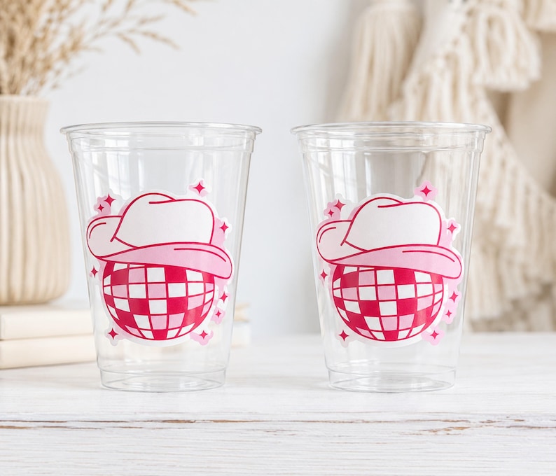 May include: Two clear plastic cups featuring a pink and white graphic of a cowboy hat over a disco ball. The disco ball has a red and white checkered pattern. Pink star accents surround the graphic. The cups are on a white wooden surface.