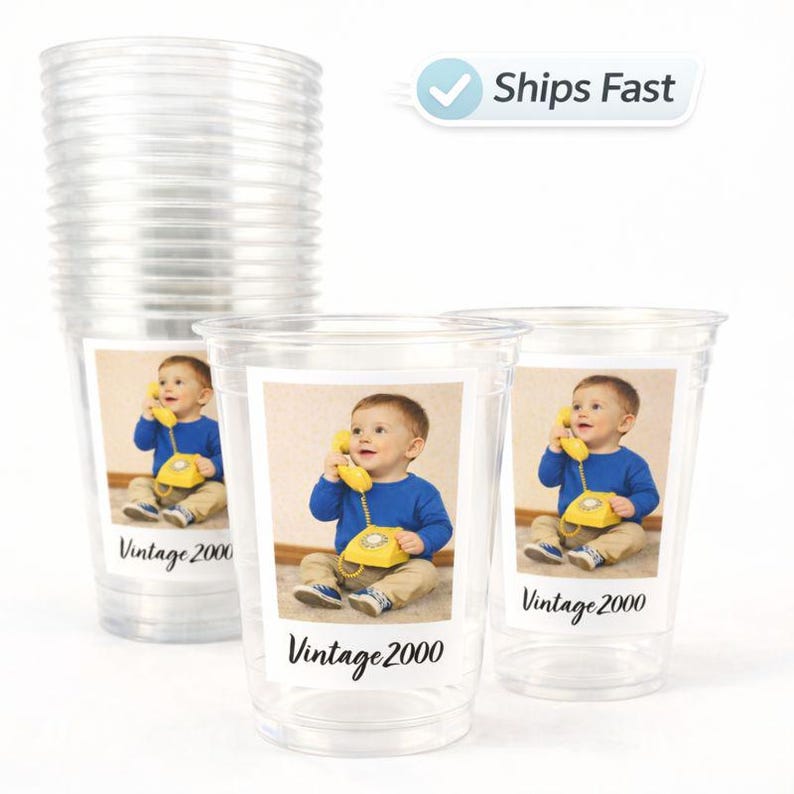May include: Clear plastic cups with a printed label featuring a baby holding a yellow telephone. The label also includes the text "Vintage 2000". A stack of similar cups is visible in the background.
