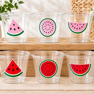 May include: A set of six clear plastic cups arranged in two rows. The top row features three cups with light pink watermelon graphics, while the bottom row features three cups with vibrant red watermelon graphics in slice and round shapes.