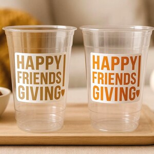May include: Two clear plastic cups with the words "HAPPY FRIENDS GIVING" in gold and orange lettering. The cups are on a wooden tray, with a small bowl of nuts and a decorative pumpkin in the background. The theme is Thanksgiving.