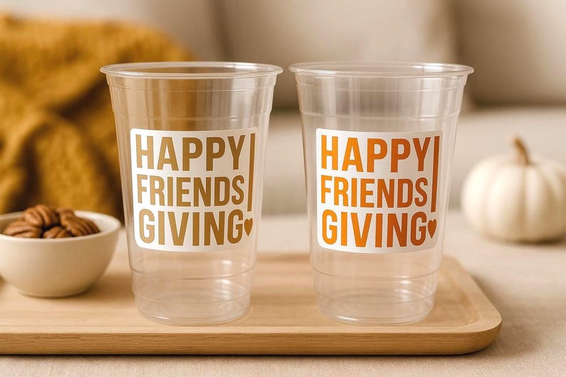 May include: Two clear plastic cups with the words "HAPPY FRIENDS GIVING" in gold and orange lettering. The cups are on a wooden tray, with a small bowl of nuts and a decorative pumpkin in the background. The theme is Thanksgiving.