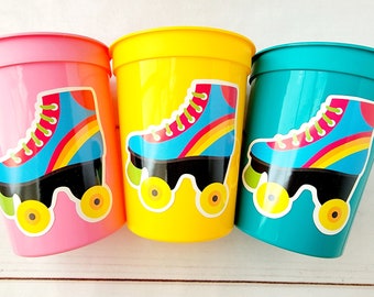 Puede incluir: Tres vasos de plástico con diseños de patines de colores. Los vasos son rosa, amarillo y azul verdoso.