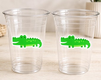 Może przedstawiać: Dwa przezroczyste plastikowe kubki z zieloną naklejką z aligatorem. Aligator ma zielone ciało, biały brzuch i czarne oczy. Kubki są puste i wydają się jednorazowe.
