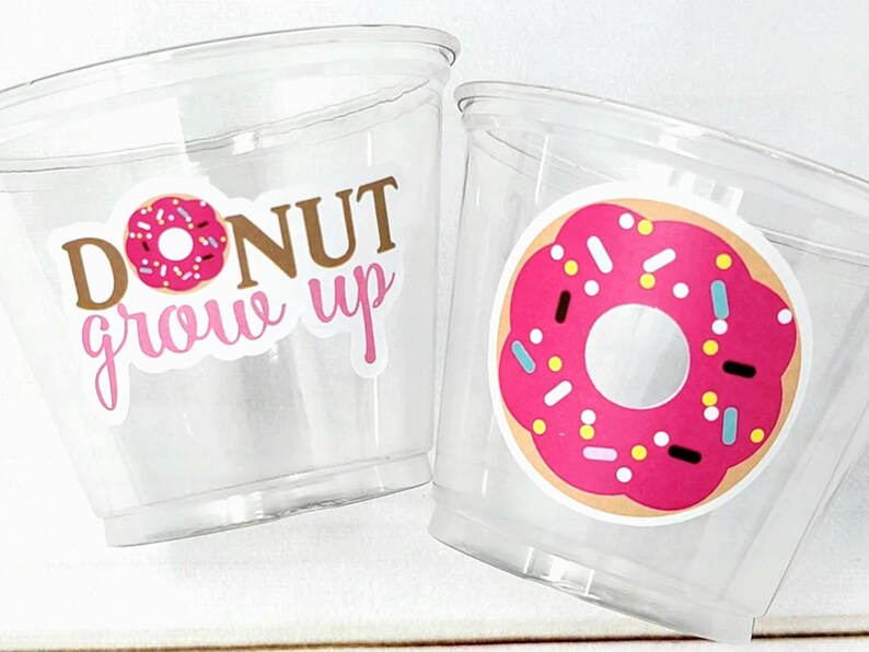 May include: Two clear plastic cups with donut-themed designs. One cup has a pink donut with sprinkles and the text "DONUT grow up". The other cup has a pink donut with sprinkles.