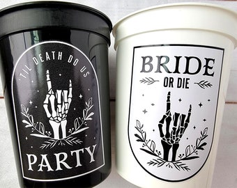 May include: A black plastic cup with white text reading 'TIL DEATH DO US PARTY' and a skeleton hand graphic, alongside a white plastic cup with black text reading 'BRIDE OR DIE' and a matching skeleton hand graphic, both featuring floral accents.
