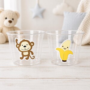 May include: Two clear plastic cups with cartoon stickers. One cup has a brown monkey sticker, and the other has a yellow banana sticker. The cups are on a white wooden surface, with a teddy bear and pillows in the background.