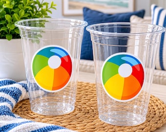 May include: Two clear plastic disposable cups, each featuring a circular sticker with a colorful beach ball graphic. The beach ball design consists of segments in blue, green, yellow, orange, and red, centered around a white circle.