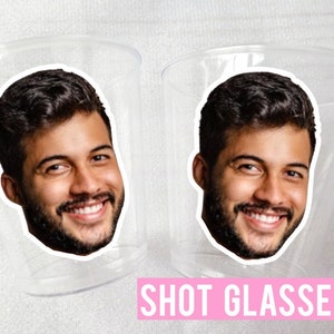 May include: Two clear plastic shot glasses with a printed image of a man's face on each. The text "SHOT GLASSES" is printed in pink below the glasses.
