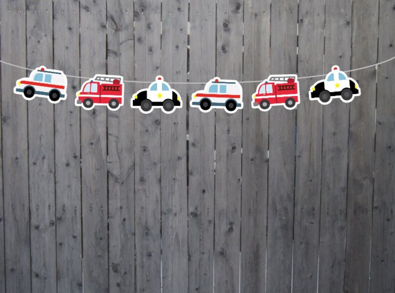 May include: A string of six paper cutouts of vehicles, alternating between red fire trucks and white police cars. The vehicles are strung together with white string and are hanging against a gray wooden fence.