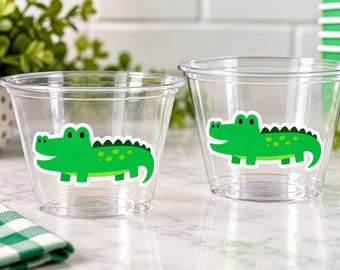 Może przedstawiać: Dwa przezroczyste plastikowe kubki z grafiką jaskrawozielonego krokodyla z kreskówki z przodu. Każdy kubek ma szeroki brzeg i zwężaną podstawę, zaprojektowane do serwowania napojów lub przekąsek podczas różnych wydarzeń.