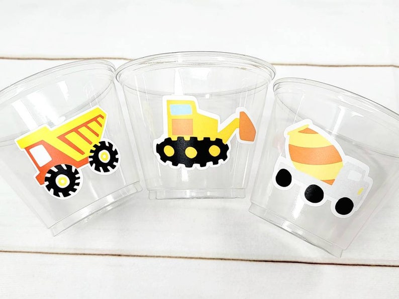 May include: Three clear plastic cups with construction vehicle designs. The cups feature a yellow dump truck, a yellow cement mixer, and a yellow excavator.