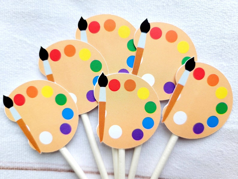 May include: Six colourful paint palette cupcake toppers with brown paintbrushes. The palettes are decorated with circles of different colours, including red, orange, yellow, green, blue, and purple.