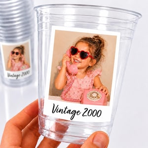 May include: A clear plastic disposable cup featuring a rectangular sticker with a photograph of a child wearing heart-shaped sunglasses and holding a pink rotary telephone. The text Vintage 2000 is printed in black script at the bottom of the sticker.