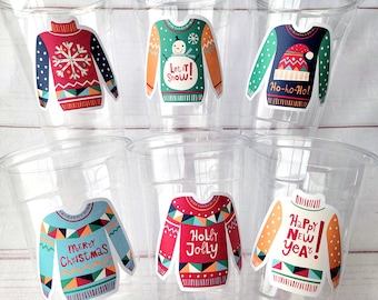 May include: Clear plastic cups decorated with colorful sweater stickers. The stickers feature holiday designs with text like "Let it Snow," "Ho-ho-Ho!," "Merry Christmas," "Holly Jolly," and "Happy New Year!" The sweaters have snowflake, snowman, and festive patterns.
