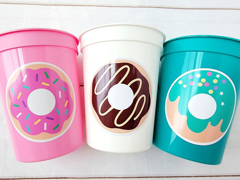 Pode incluir: Tr&ecirc;s copos de pl&aacute;stico com desenhos de donuts. Os copos s&atilde;o rosa, branco e azul turquesa. Os donuts t&ecirc;m cobertura rosa, chocolate e verde com granulado.