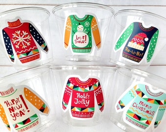 May include: Clear plastic cups decorated with colorful stickers featuring holiday sweater designs. The stickers include phrases like "Let it Snow!", "Ho-ho-Ho!", "Happy New Year!", "Holly Jolly", and "Merry Christmas".