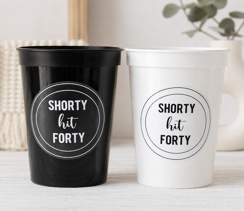 May include: Two plastic cups, one black and one white, each with a circular sticker. The stickers read "SHORTY hit FORTY" in a black and white design. The cups are likely for a birthday celebration.