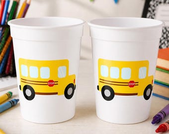 Op de afbeelding: Twee witte plastic bekers, elk voorzien van een grafische illustratie van een gele schoolbus op de zijkant. Het busontwerp bevat lichtblauwe ramen, zwarte wielen en een rood stopbord. De bekers zijn glad, cilindervormig en hebben een brede rand.