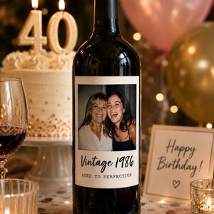 May include: A dark glass wine bottle with a custom rectangular label featuring a photograph of two smiling individuals. The label includes the printed text Vintage 1986 and AGED TO PERFECTION in black lettering against a cream-colored background.
