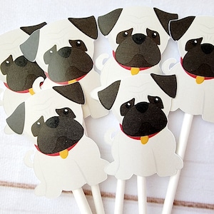 May include: Six white paper cupcake toppers shaped like pugs with black faces, red collars, and yellow name tags. The toppers are on white sticks.