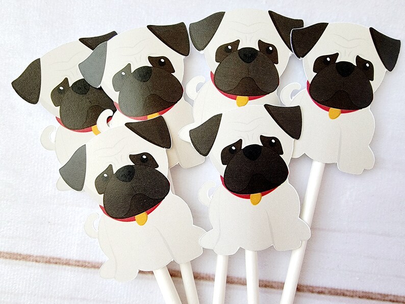 May include: Six white paper cupcake toppers shaped like pugs with black faces, red collars, and yellow name tags. The toppers are on white sticks.