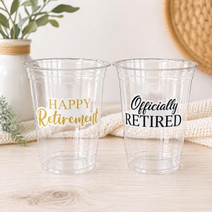 May include: Two clear plastic cups with the phrases "Happy Retirement" and "Officially Retired" in gold and black lettering. The cups are on a light wooden surface, with a vase of greenery and a woven basket in the background.