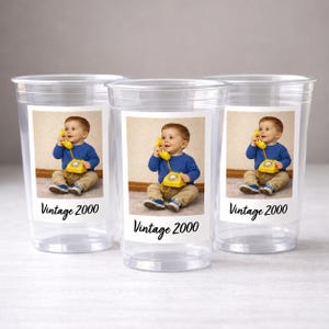May include: Three clear plastic cups with a printed label featuring a baby holding a yellow vintage telephone. The label also includes the text "Vintage 2000". The baby is wearing a blue shirt and khaki pants.