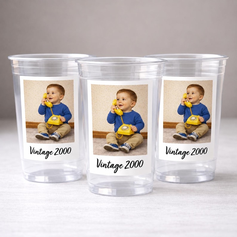 May include: Three clear plastic cups with a printed label featuring a baby holding a yellow vintage telephone. The label also includes the text "Vintage 2000". The baby is wearing a blue shirt and khaki pants.