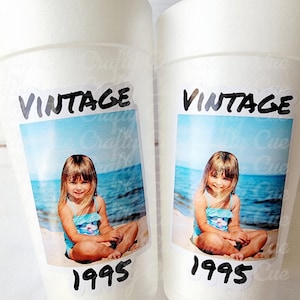 May include: Two white foam cups with a vintage photo sticker. The sticker features a photo of a young girl in a blue swimsuit on a beach, with the word "VINTAGE" above and "1995" below.