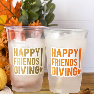 May include: Two clear plastic cups with the words "HAPPY FRIENDS GIVING" in gold and orange lettering. The cups are filled with a white liquid and are decorated with a heart. The background includes fall foliage and cinnamon sticks.