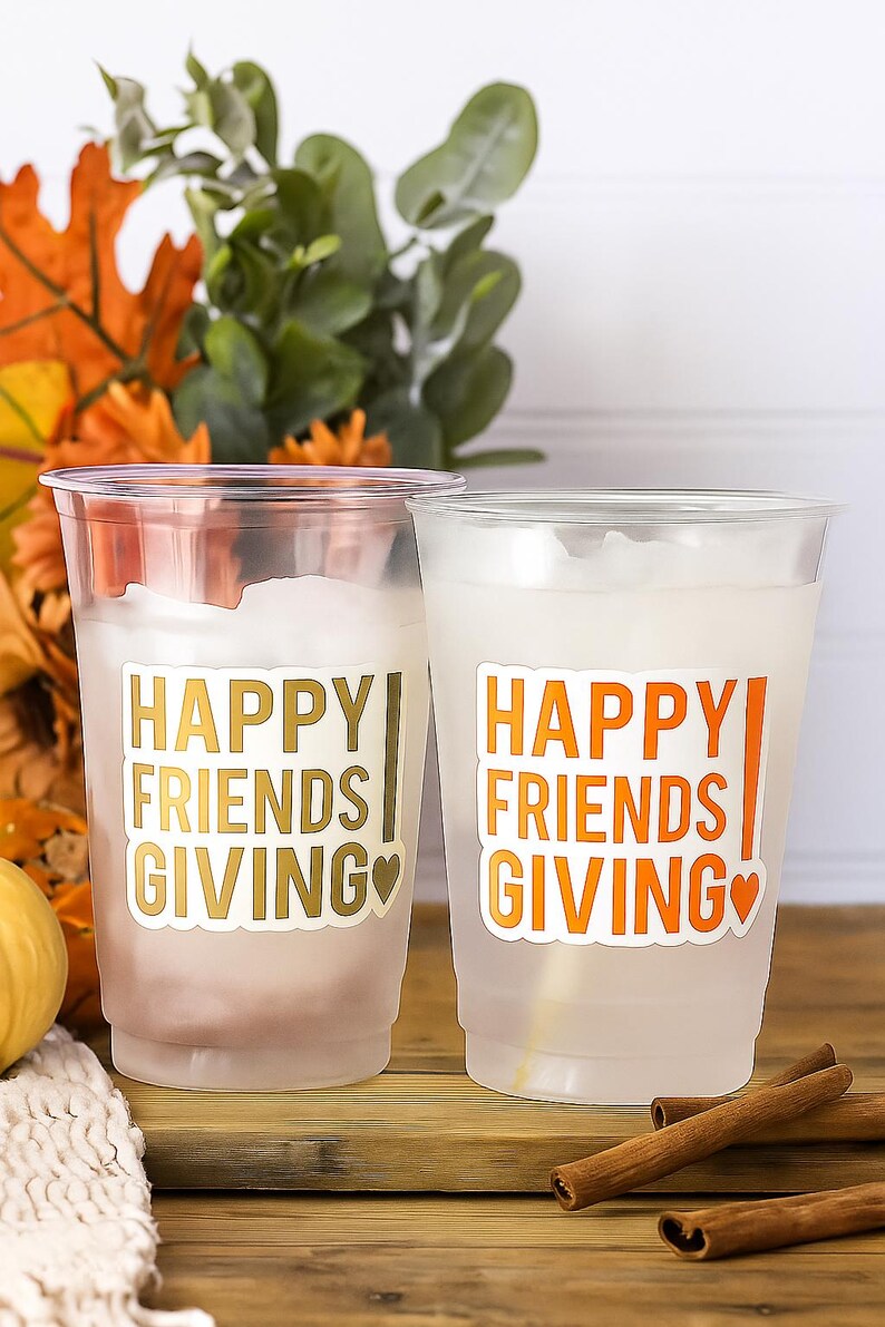 May include: Two clear plastic cups with the words "HAPPY FRIENDS GIVING" in gold and orange lettering. The cups are filled with a white liquid and are decorated with a heart. The background includes fall foliage and cinnamon sticks.