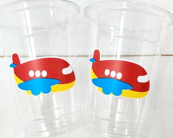Puede incluir: Dos vasos de plástico transparentes con pegatinas de avión rojo, amarillo y azul. Los aviones tienen tres círculos blancos en las alas.