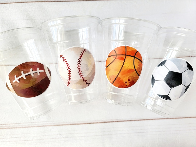 May include: Four clear plastic cups with different sports ball designs. The cups have a brown football, a white baseball, an orange basketball, and a black and white soccer ball.