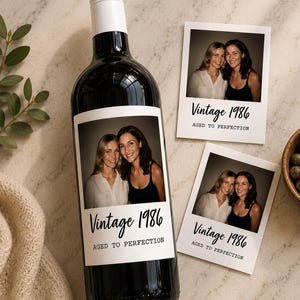 May include: A dark glass wine bottle with a white label featuring a photograph of two people and the text Vintage 1986 AGED TO PERFECTION. Two matching square photo prints with the same text are placed next to the bottle on a light surface.