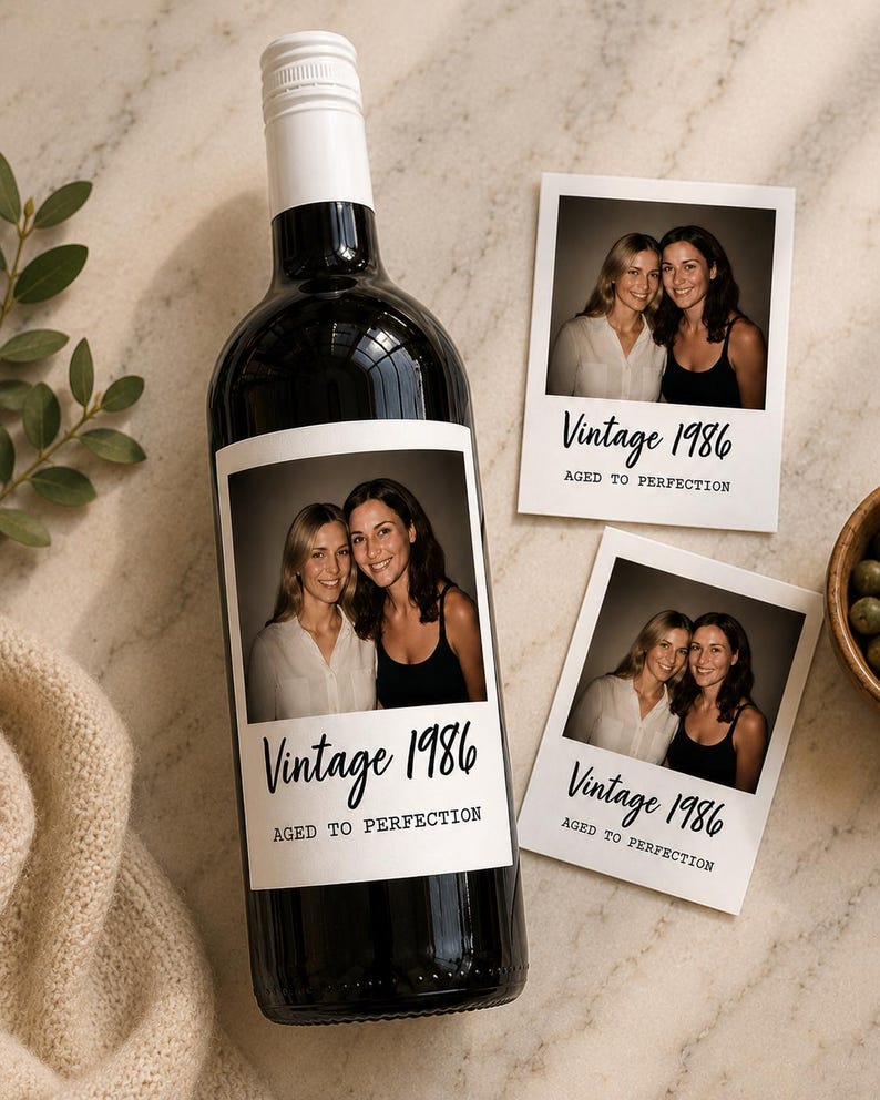 May include: A dark glass wine bottle with a white label featuring a photograph of two people and the text Vintage 1986 AGED TO PERFECTION. Two matching square photo prints with the same text are placed next to the bottle on a light surface.