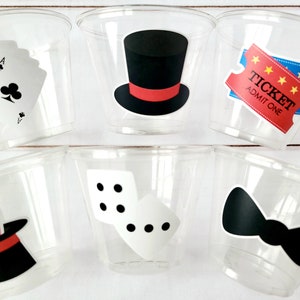 May include: Six clear plastic cups with different magician-themed designs. The designs include a top hat, playing cards, a ticket, dice, a bow tie, and a top hat with bunny ears.